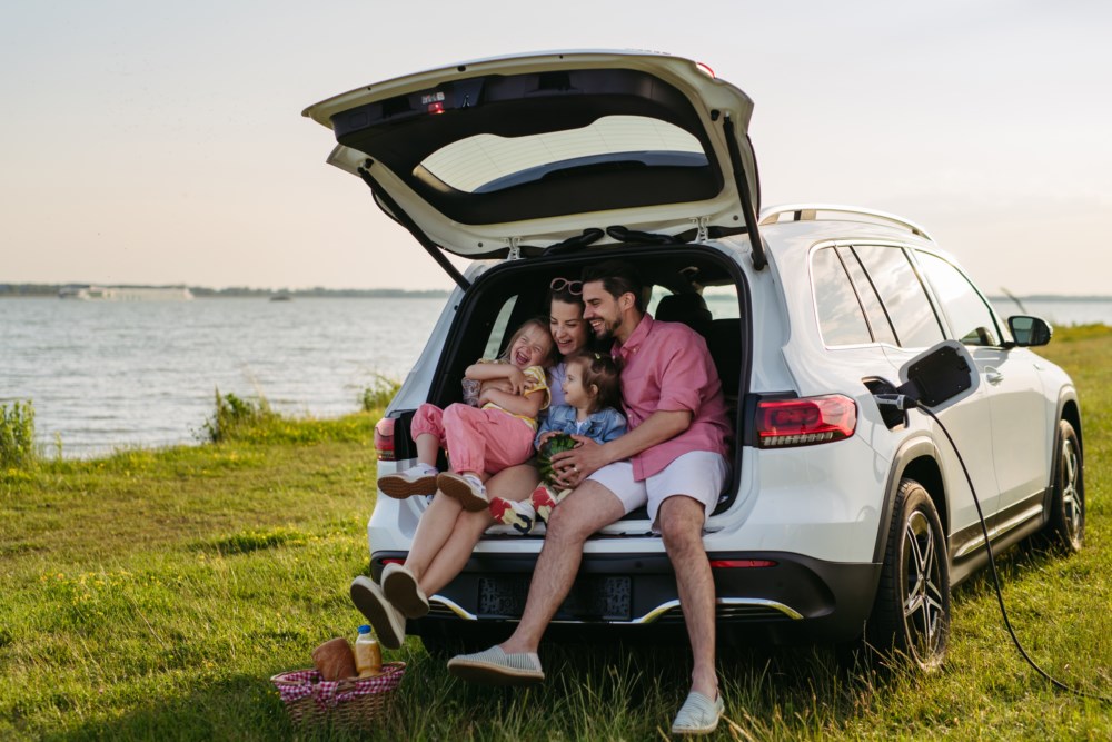 Familie på fire koser seg i bagasjerommet på bilen, parkert ved en innsjø med en piknikkurv ved siden av.